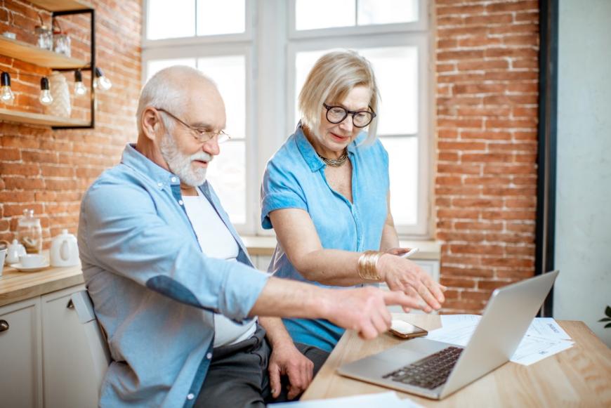 Older man and woman in the kitchen and pointing at their laptop.