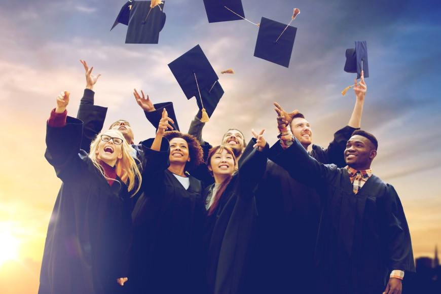 Multiracial group of college grads in graduation gowns celebating and throwing their mortar boards in the air