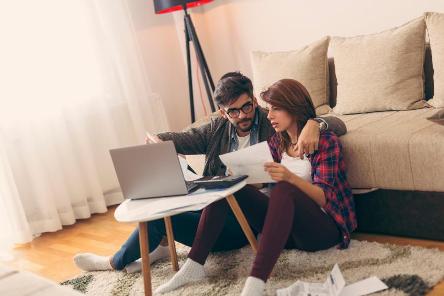 Couple sitting on floor in front of couch, using a laptop and looking at papers