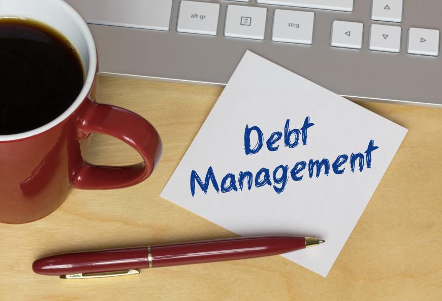 Post it that says Debt Management and resting on desk.  Next to note are a cup of coffee, a computer keyboard, and a red pen.