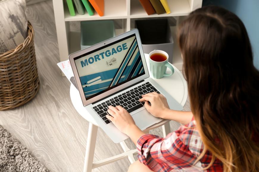 Woman with long reddish hair typing on her laptop. The top of laptop displays the word Mortgage.