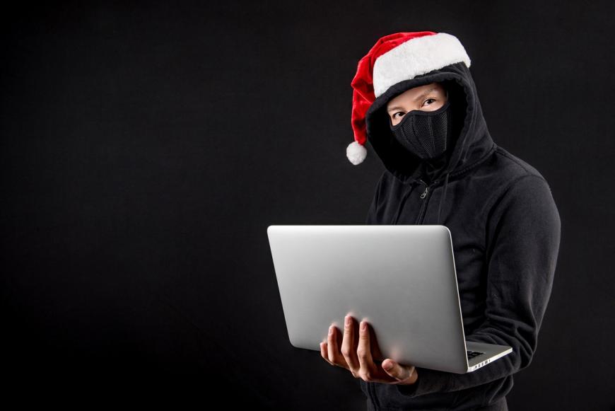 Man wearing a Santa hat and a black sweater with a black face mask and holding a laptop.
