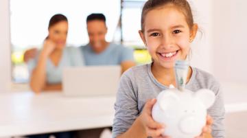 Young girl holding piggy bank and smiling.  A couple in the background looking at information their laptop.