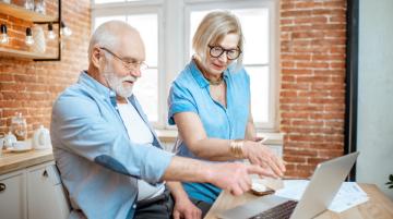 Older man and woman in the kitchen and pointing at their laptop.