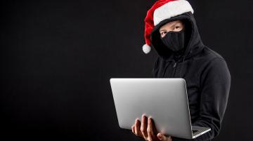 Man wearing a Santa hat and a black sweater with a black face mask and holding a laptop.