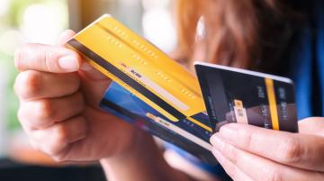 Person holding three credit cards (one black, one yellow, and one blue) with the left hand and pulling the yellow card from the pile with the right hand.