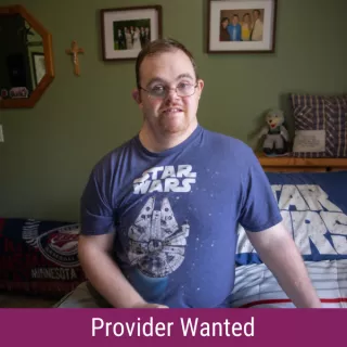 A man with Down syndrome sitting on a bed, wearing a Star Wars t-shirt and smiling, with the text "Provider Wanted"